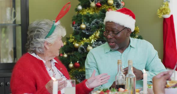 Happy diverse senior couple in santa hats talking at christmas dinner table at home