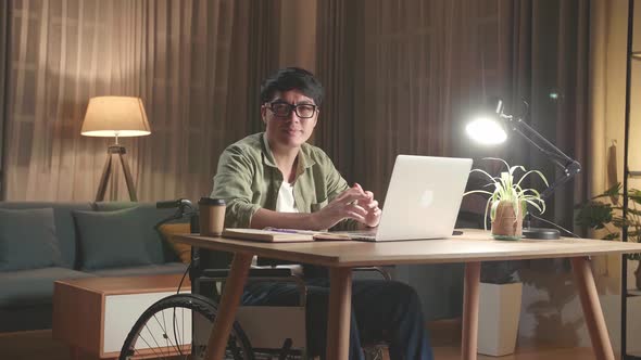 Asian Man Sitting In A Wheelchair Working On Laptop Computer While Looking And Smiling To Camera