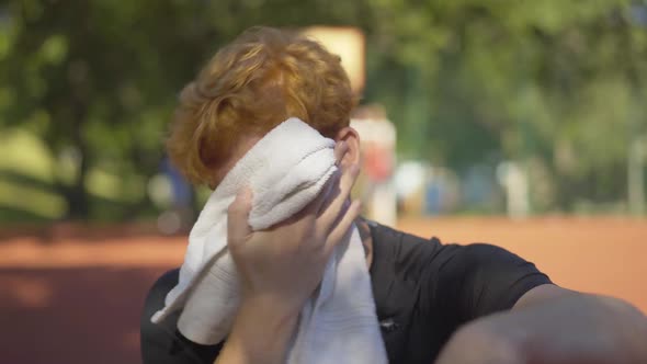 Close-up of Perspiring Young Redhead Sportsman Wiping Face with White Towel Outdoors. Portrait of