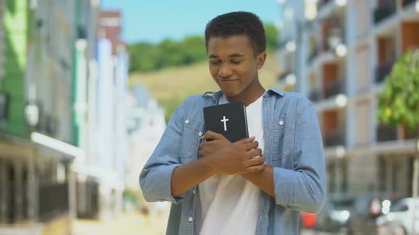 Glad African-American Young Male Hugging Holy Bible and Looking at Church, Faith