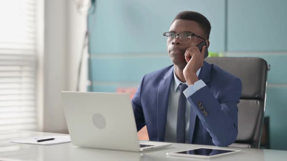 Young African Businessman Talking on Phone While Using Laptop in Office