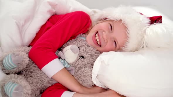 Smiling Girl in Santa Hat and Christmas Pajamas Lying in Bed with Teddy Bear at Home