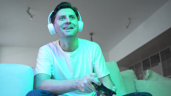 Gambling Male Gamer Playing Video Game at Home with Joystick and Headphones While Sitting on Sofa