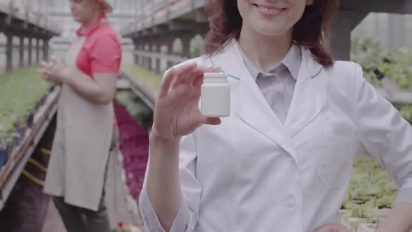 Unrecognizable Woman Showing Bottle with Pesticides at Camera As Blurred Man Examining Flower in Pot