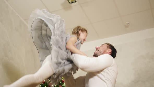 Young Father is Throwing His Adorable Daughter in the Air at Home with Decorated Christmas Tree on