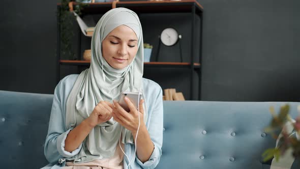 Young Muslimah Speaking on Mobile Phone Using Earphones with Microphone Smiling Indoors at Home