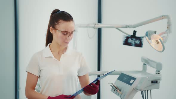 Caucasian Female Dentist Doctor in Dental Office in Protective Transparent Mask and Folder in Hands