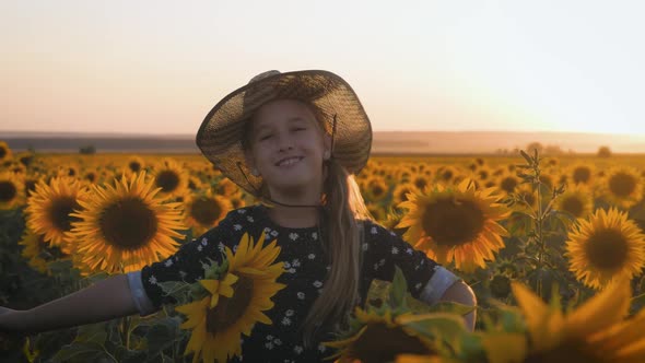 A Happy Little Girl in the Hat Is Smiling at the Field of Sunflowers at Sunset Time