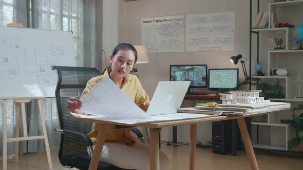 Asian Woman Engineer Looking At Blueprint And Typing On A Laptop At The Office