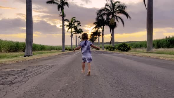 A Little Girl in a Striped Dress Runs Along the Road with Palm Trees Against the Background of a