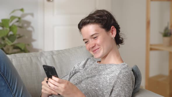 Young Woman Relax on Couch with Smartphone Spend Free Time at Home Looks at Cell Phone Screen Typing