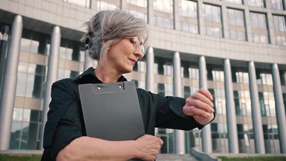 Adult Serious Lady Waits Near a Business Office Looks at Her Watch and Holds a Folder with Documents