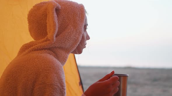 Woman Traveler in Funny Bunny Pajama Enjoying Hot Tea In Mug On Beach in Tent at Cold Morning