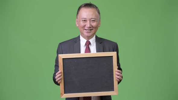 Mature Japanese Businessman Holding Blackboard Against Green Background