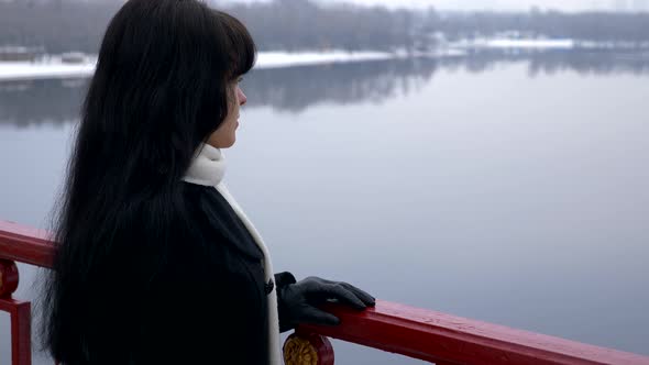 Beautiful Brunette Stands On Bridge Above River Thinking And Looking Ahead. Slow Motion Winter