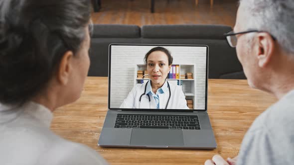 Back View of Aged Couple Looking at Screen of Laptop and Talking to Doctor Woman Using Online Video