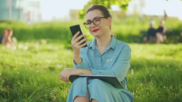 Woman Using Smartphone While Sitting in Park After Finishing Outdoor Work at Sunset