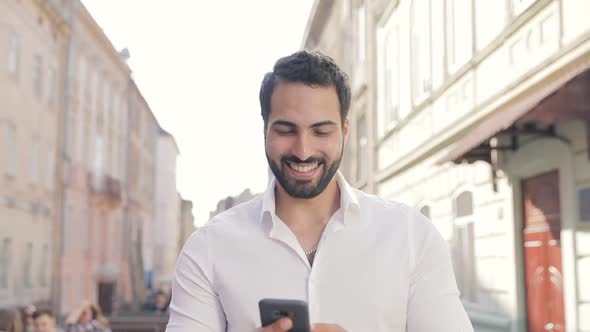 Smiling Man Using Mobile Phone And Walking Along City Street