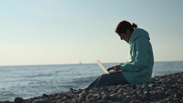 Caucasian Woman Typing on a Laptop on a Rocky Seashore