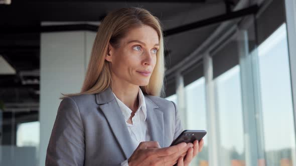 Young Positive Woman is Standing Near Window in Office Browsing Her Smartphone