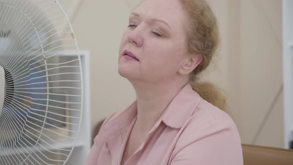 Close-up of Senior Woman Using Electric Fan Indoors. Portrait of Ill Caucasian Lady Having Fever or