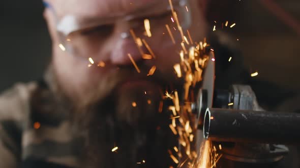 Bearded Male Craftsman Works in Workshop Cutting Metal Pipe From Which Sparks Fly Uses an Angle