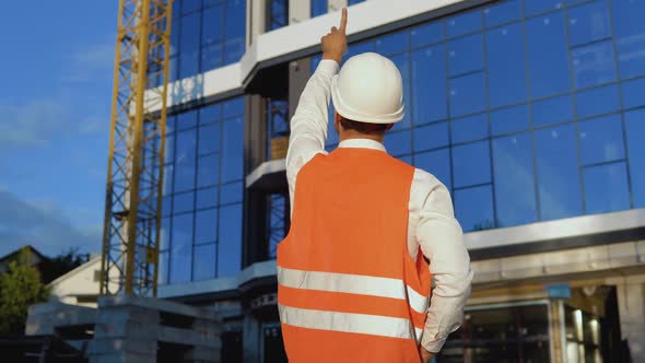 An Engineerarchitect in a White Shirt Helmet and Orange Work Vest Stands with His Back to the Camera