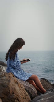 A Beautiful Young Woman is Reading a Book Sitting on a Large Stone on Seashore