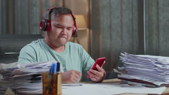 Fat Asian Man Wearing Headphones And Listening To Music On Smartphone After Working With Documents