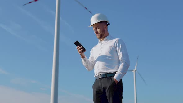 An Engineer in Business Clothes and Helmet Works in a Cell Phone Against the Background of Wind