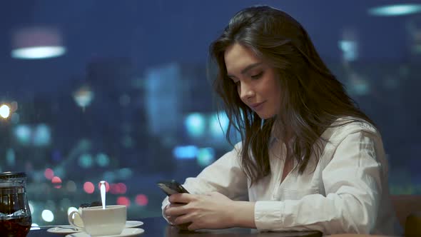 Young Girl Is Texting a Message on the Smartphone While Sitting in a Cafe on the Background of the