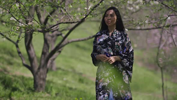 Young Slim Charming Asian Woman in Kimono Walking in Sunny Spring Park Turning and Smiling Looking