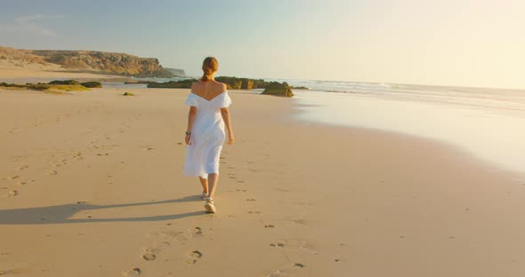 Woman in a White Dress Walks Along Sandy Golden Beach Towards the Sea Against the Backdrop of a