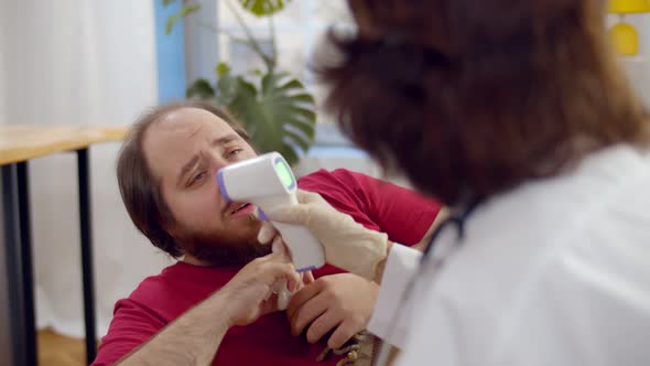 Doctor in Protective Mask Measures Temperature with Noncontact Thermometer To Patient at Home