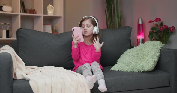 Little Joyful Girl in Headphones is Dance on Sofa and Using Smartphone