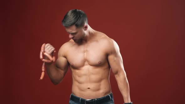 Muscular bodybuilder on dark red background. Athlete with naked torso lifting sausage