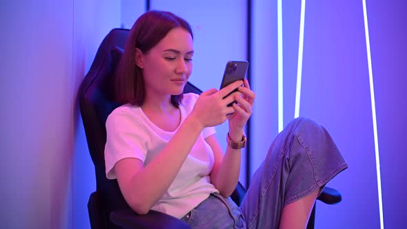 Young Lady Sitting on Gaming Chair with Cellphone in Hands. Playing Online Mobile Game or Browsing