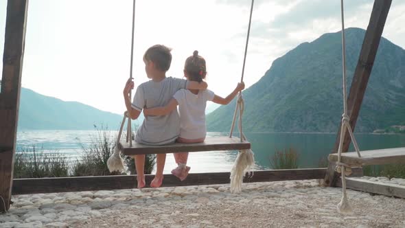 A Girl and a Boy Swing on a Swing Near the Sea with a Beautiful View of the Bay and Mountains