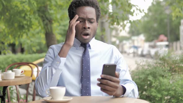 African Businessman Upset by Loss on Smartphone, Sitting in Outdoor Cafe