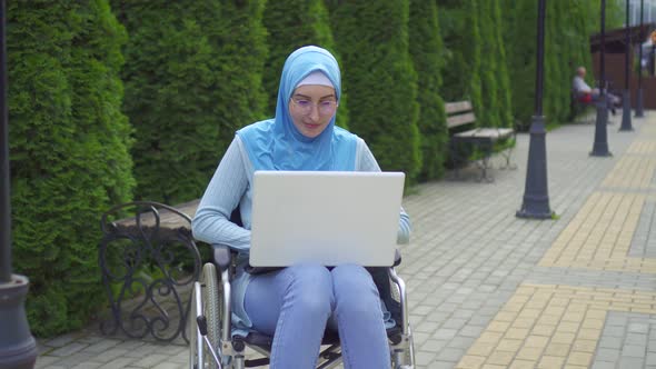 Portrait Young Muslim Woman Disabled in a Traditional Scarf Uses a Laptop Sitting in a Wheelchair