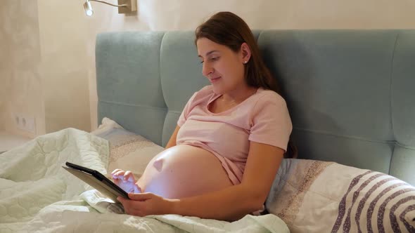 Portrait of Young Pregnant Woman with Big Belly Lying in Bed with Tablet Computer and Browsing