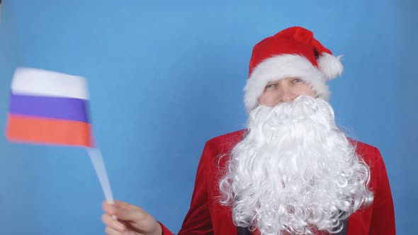 A Man in a Santa Claus Costume with a Beard Waving the Flag of Russia on a Blue Background