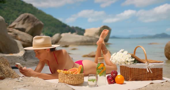 Lovely Woman in Bikini and Hat is Lying with Legs Raised and Texting on a Picnic on Isolated Beach