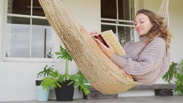 Smiling caucasian woman lying in hammock reading book on terrace