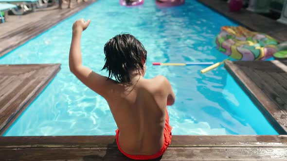 Back View of Playful Caucasian Boy Having Fun Dancing Sitting on Poolside in Sunshine