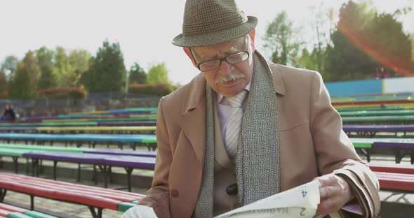 Portrait of Old Grandfather Reading Newspaper on Bench Alone in Sunny Park