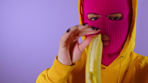 Close Up of Young Woman in Pink Balaclava and Yellow Hood Peeling Banana on Purple Background