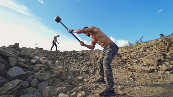 Athletic men doing hard exercises with stones. Shirtless sportsman breaks rocks with a sledgehammer 