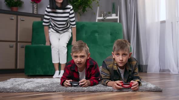 Woman Passing by Her Handsome Positive Smiling Sons which Lying on the Floor and Playing Video Game