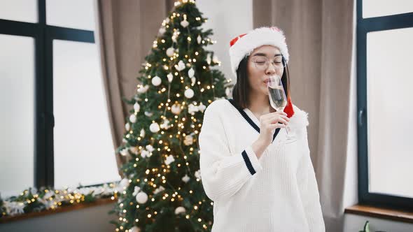 Chinese Female in Santa Hat is Drinking Champagne and Smiling While Standing at Home Against